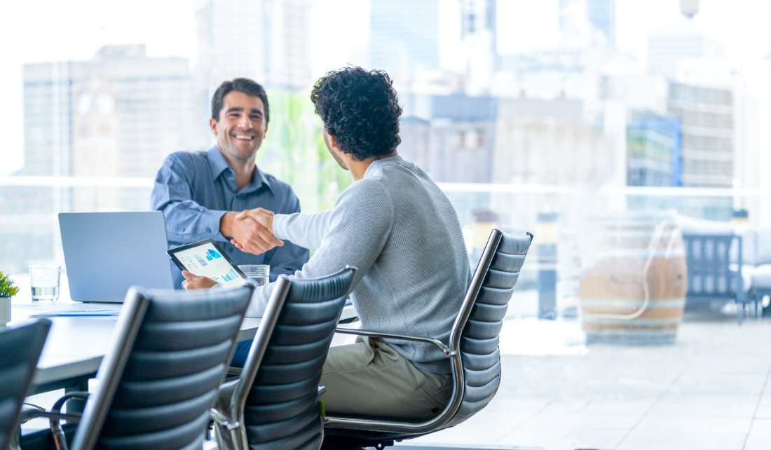 Two businessmen shaking hands in a bright office. Laptop and tablet with charts on the table. Cityscape visible through the window.